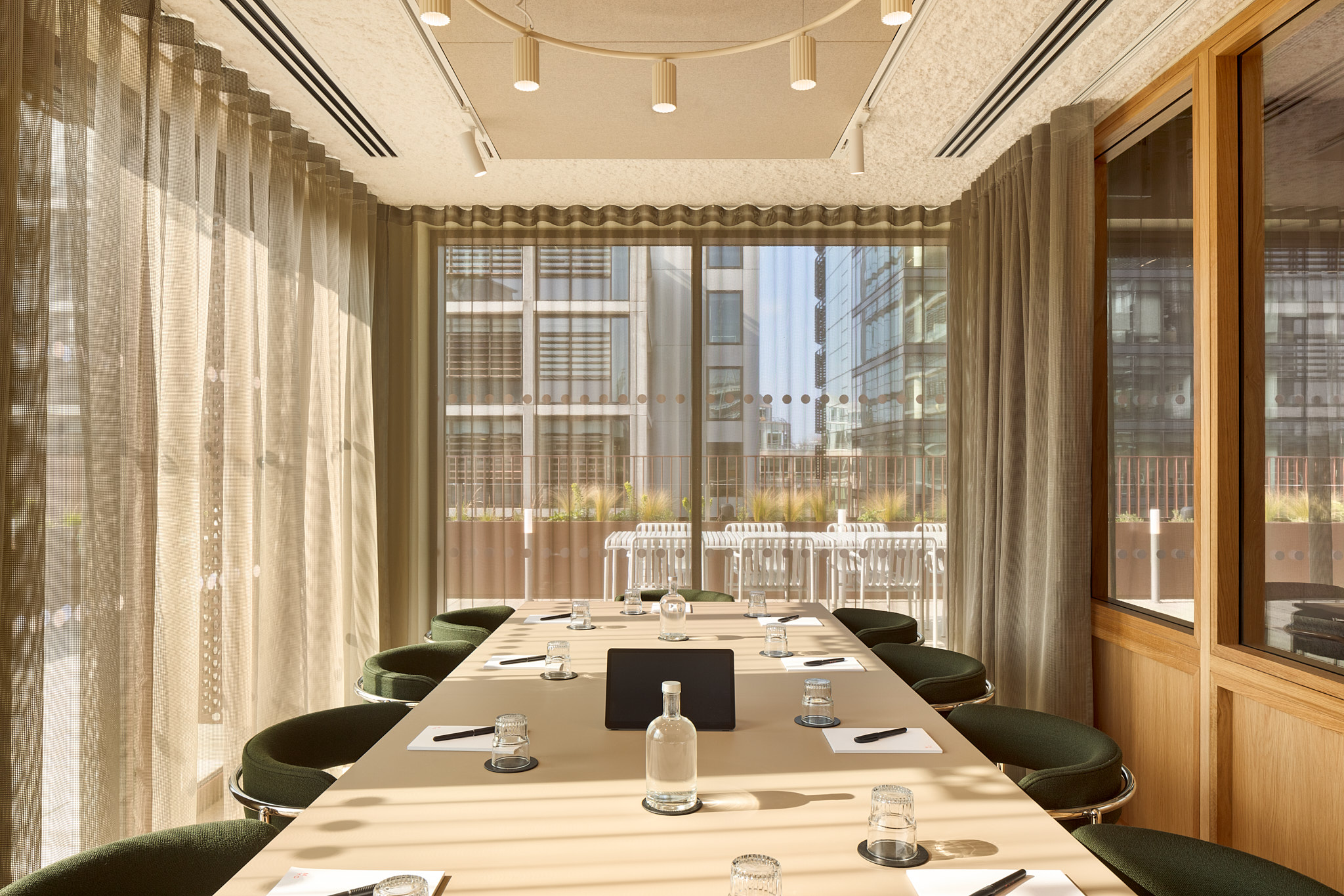 A contemporary meeting room featuring a conference table, upholstered chairs, floor-to-ceiling windows, sheer curtains and views of an outdoor terrace.