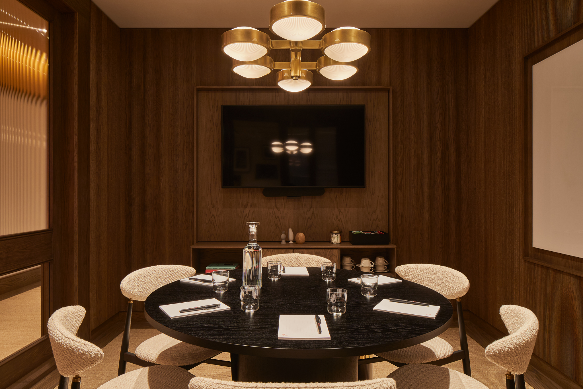 A contemporary meeting room featuring a round conference table, upholstered chairs, a wall-mounted presentation screen, wood panelling and pendant lighting.
