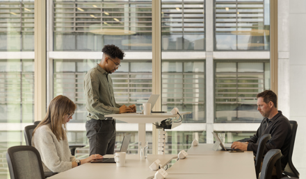 Shared workspace at MYO New Street Square featuring individuals working at ergonomic desks and standing-height workstations positioned beside floor-to-ceiling windows.