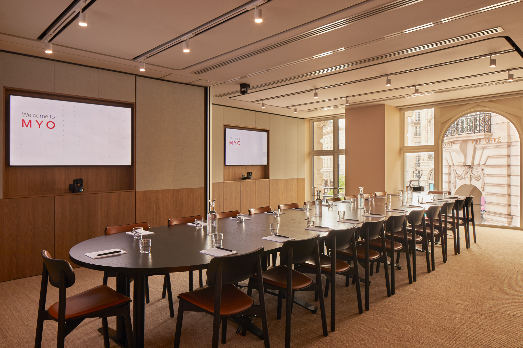 A contemporary executive boardroom featuring a long conference table, presentation screens, wooden chairs and large windows overlooking the city.