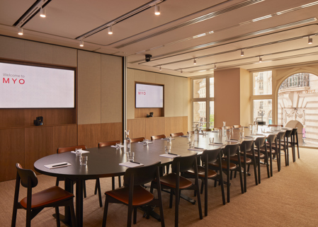A contemporary executive boardroom featuring a long conference table, presentation screens, wooden chairs and large windows overlooking the city.
