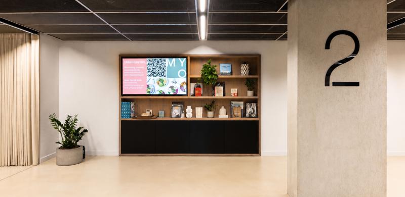 A contemporary reception area featuring a digital display, open shelving with books and plants, a large column with floor numbering and modern office interiors.