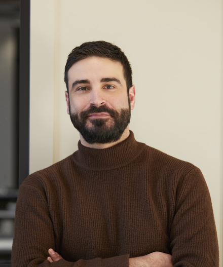 Professional headshot of a man with dark hair and a beard wearing a brown roll-neck jumper, standing with folded arms against a neutral-toned backdrop within a contemporary MYO workspace.