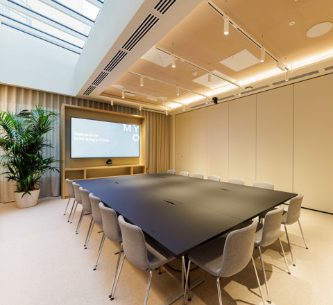 A contemporary boardroom featuring a large conference table, upholstered chairs, a presentation screen, skylight and modern lighting.