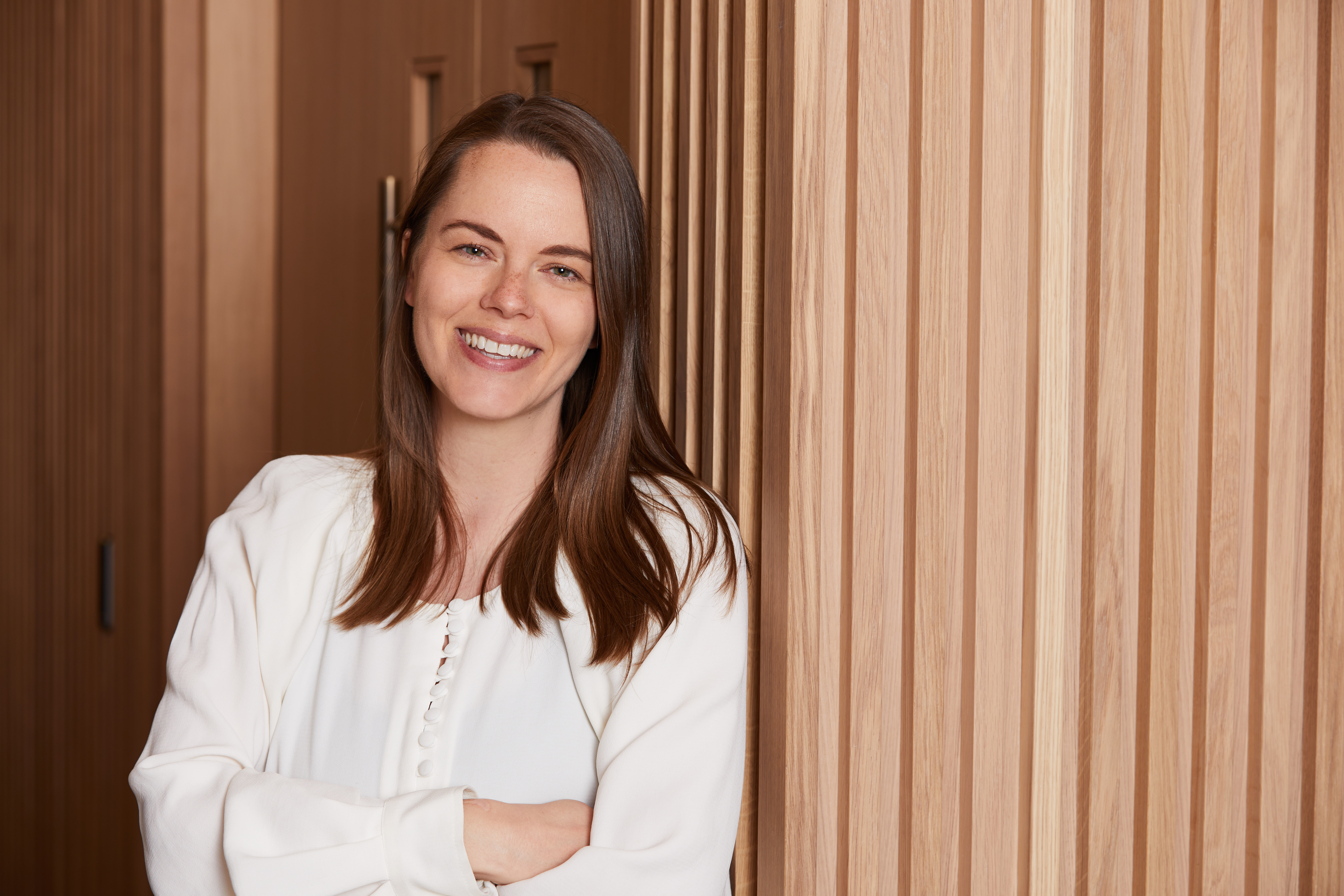 Professional headshot of a smiling woman with shoulder-length brown hair wearing a white blouse, standing beside light oak slatted wall panelling within a MYO workspace.