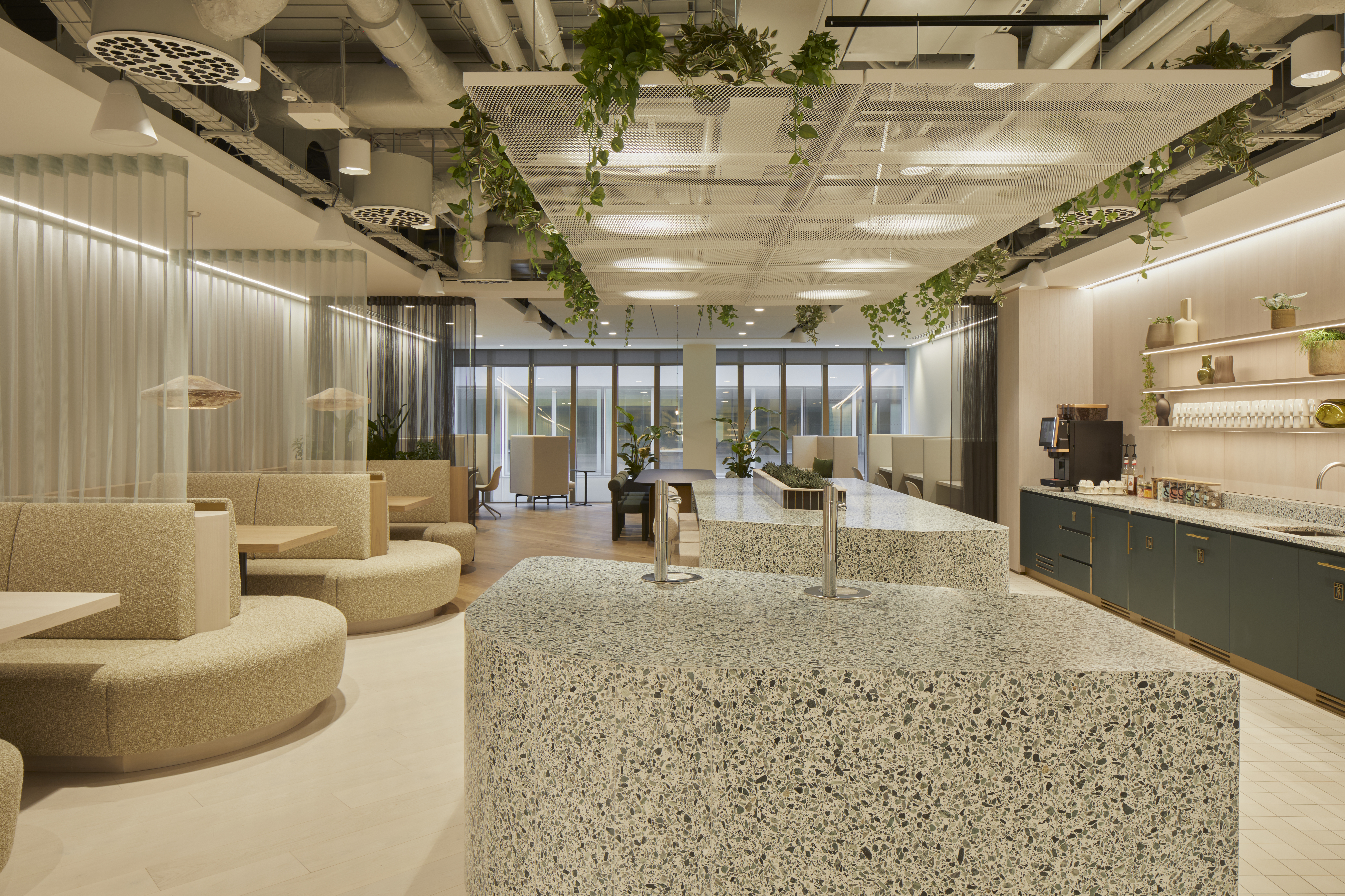 A modern kitchen and breakout area featuring terrazzo islands, curved booth seating, hanging plants, a coffee station and floor-to-ceiling windows in a contemporary workspace.