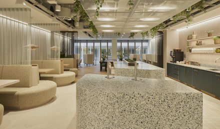 A modern kitchen and breakout area featuring terrazzo islands, curved booth seating, hanging plants, a coffee station and floor-to-ceiling windows in a contemporary workspace.