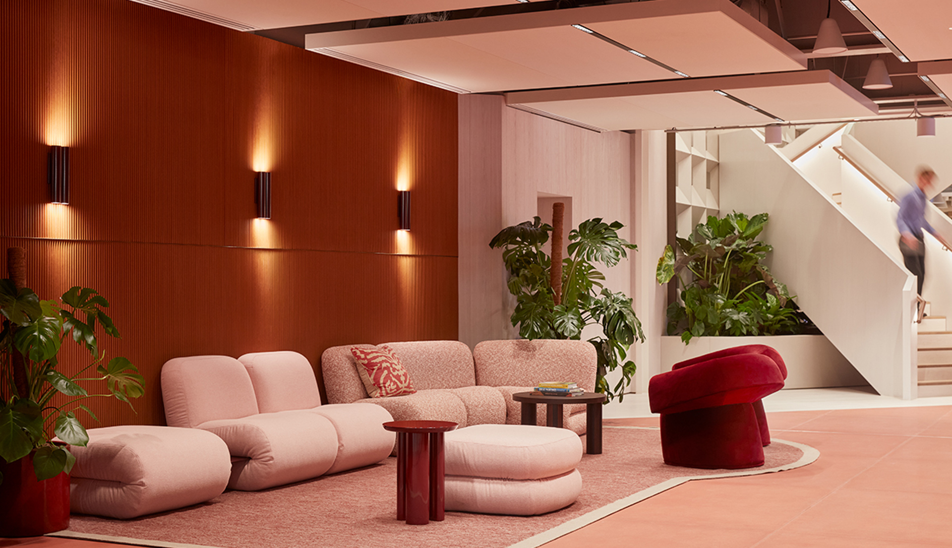 Lounge area at MYO New Street Square featuring curved blush pink seating, a statement red accent chair, rich timber wall panelling, indoor planting and an open staircase.