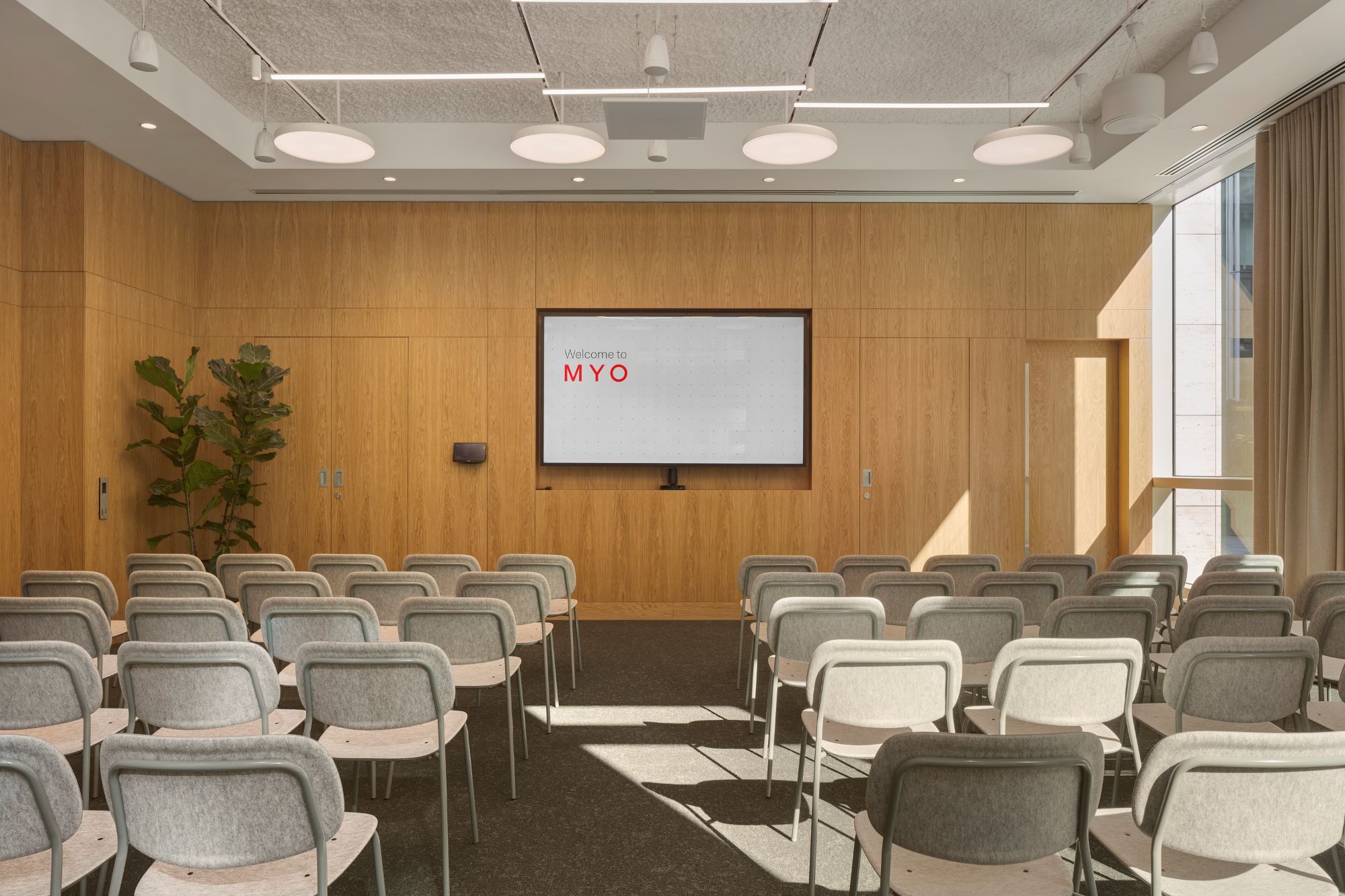A contemporary conference room with rows of upholstered chairs facing a large presentation screen, featuring timber wall panelling and floor-to-ceiling windows.