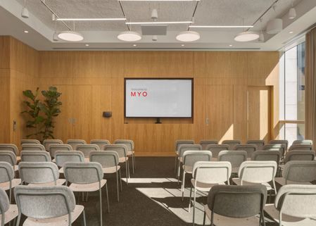 A contemporary conference room with rows of upholstered chairs facing a large presentation screen, featuring timber wall panelling and floor-to-ceiling windows.