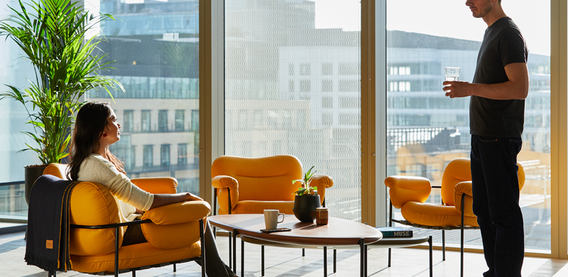 Lounge area at MYO Liverpool Street featuring mustard yellow armchairs, floor-to-ceiling windows and city views, with members enjoying an informal conversation.