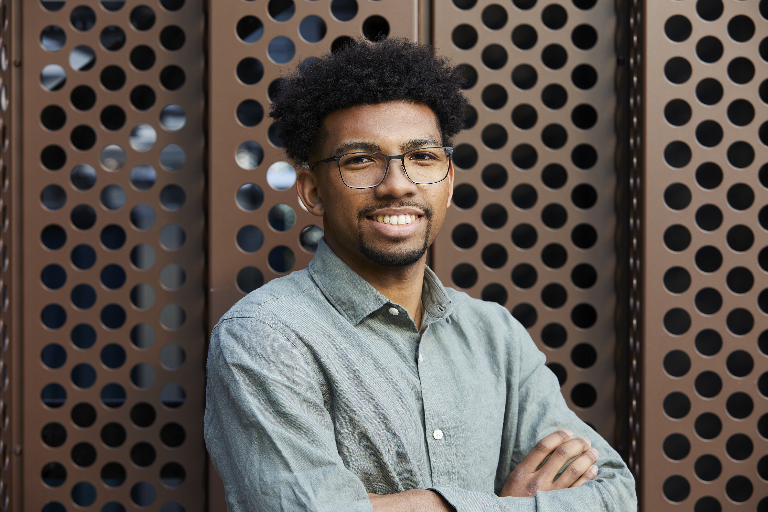 Portrait of a male MYO team member standing with arms crossed against a perforated architectural backdrop.