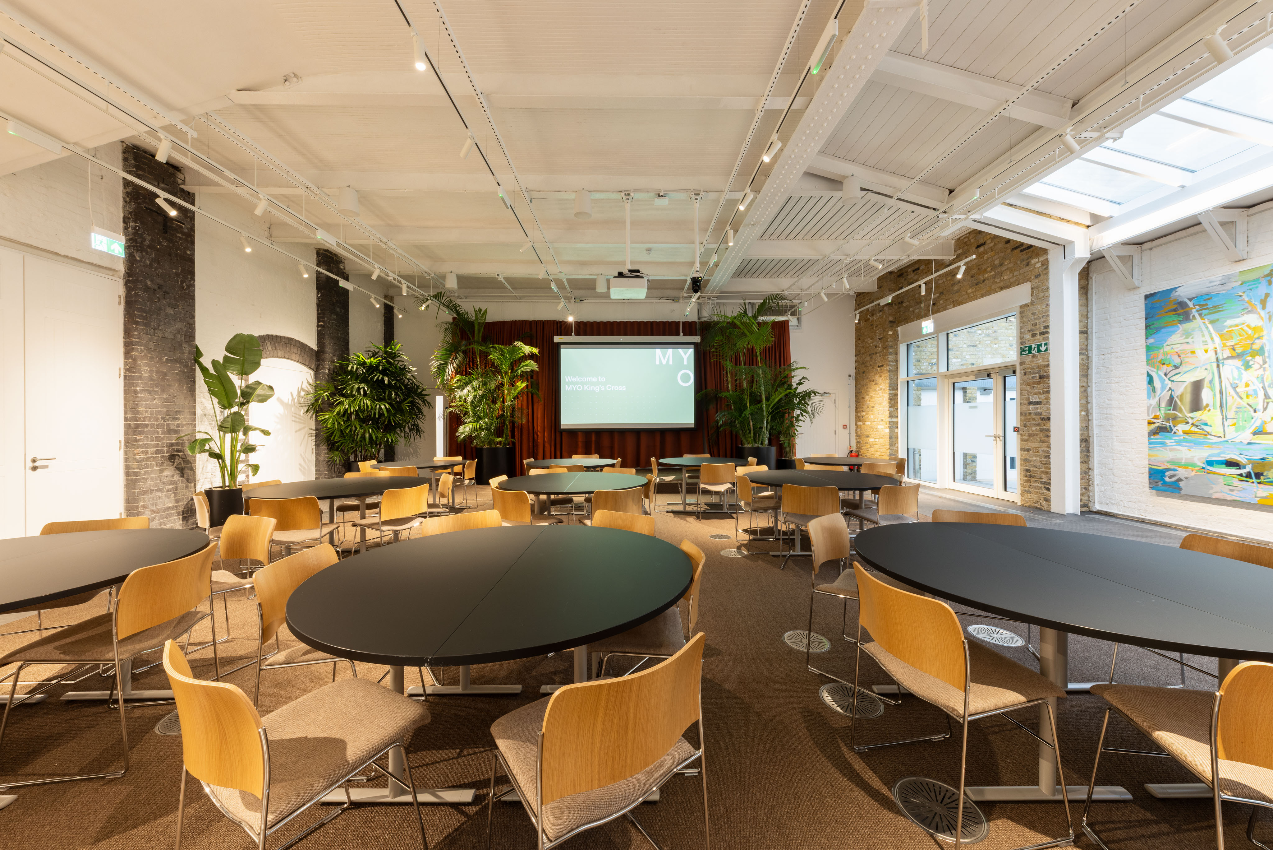 Bright event space with round black tables, wooden chairs, indoor plants and a presentation screen at the front of the room.