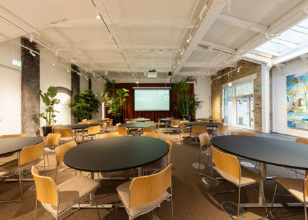 Bright event space with round black tables, wooden chairs, indoor plants and a presentation screen at the front of the room.