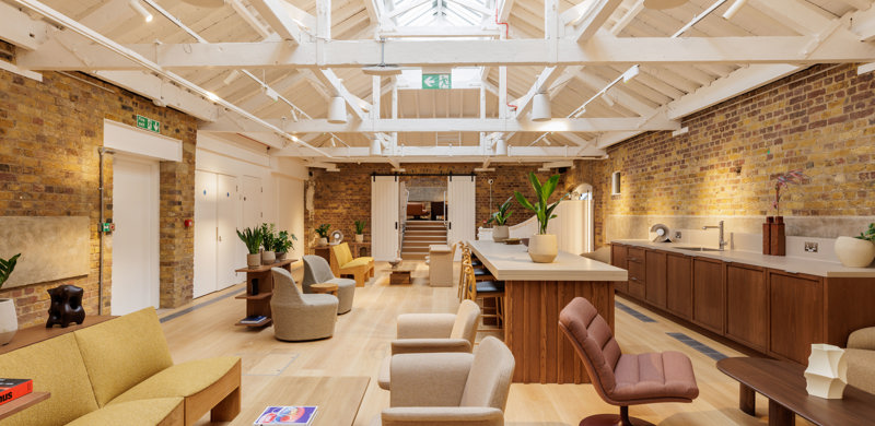 A contemporary lounge and communal kitchen featuring comfortable seating, a large kitchen island, exposed brick walls and a vaulted ceiling with skylights.