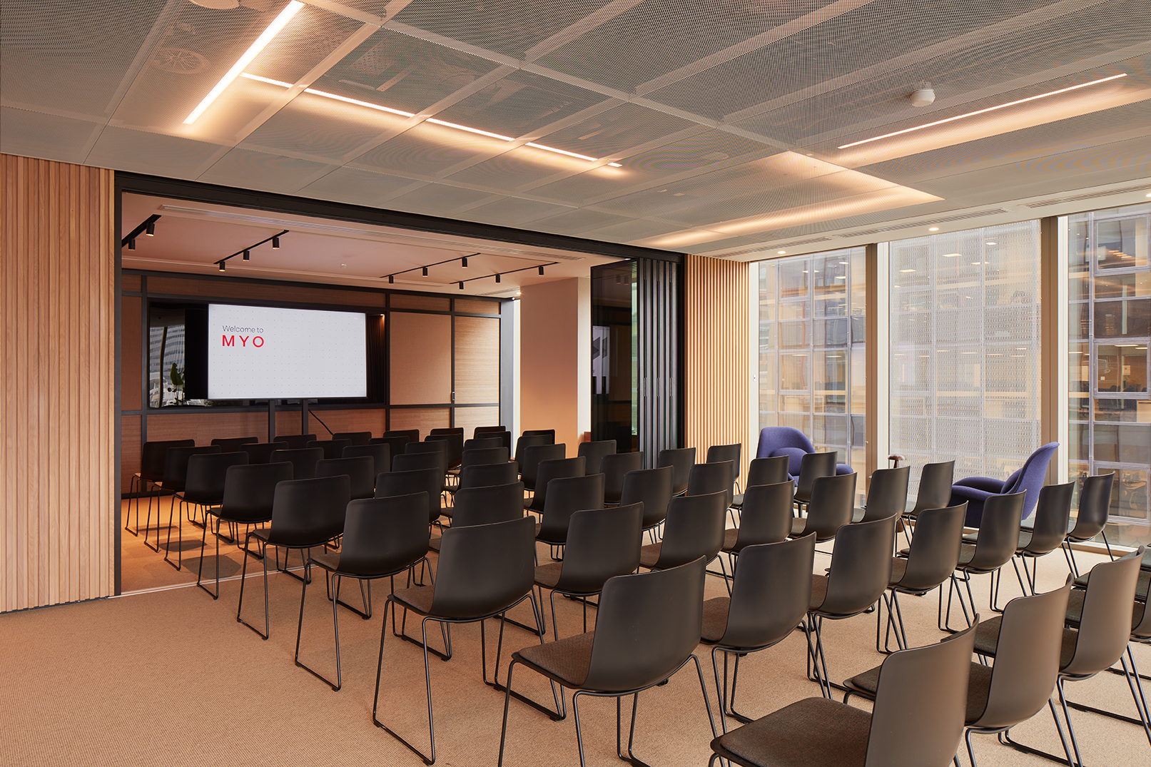 Event space at MYO Liverpool Street arranged in theatre style with rows of black chairs, presentation screen, floor-to-ceiling windows and contemporary timber detailing.