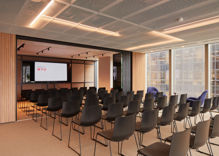Event space at MYO Liverpool Street arranged in theatre style with rows of black chairs, presentation screen, floor-to-ceiling windows and contemporary timber detailing.