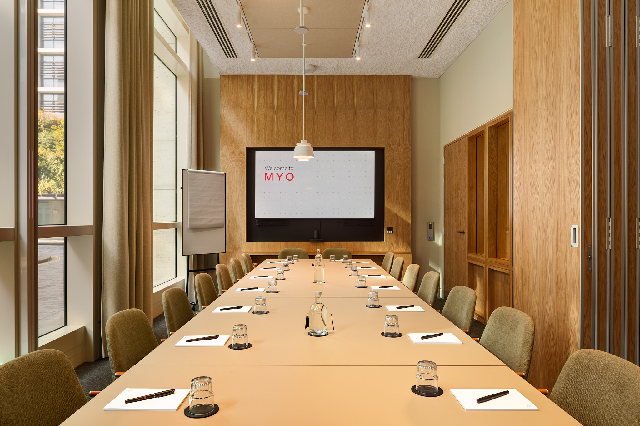 A contemporary boardroom featuring a long conference table, upholstered chairs, a presentation screen, flip chart and floor-to-ceiling windows.