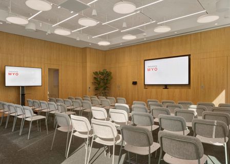 A contemporary conference room with rows of upholstered chairs, two large presentation screens, timber wall panelling and modern lighting.