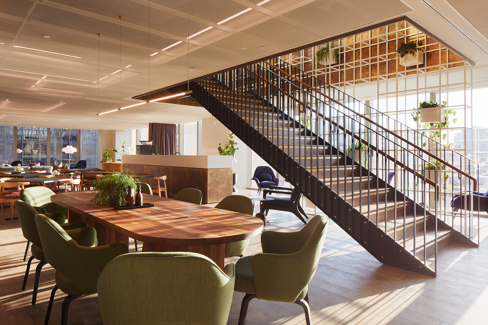 Open lounge area at MYO Liverpool Street featuring a large walnut meeting table with olive green chairs, a statement staircase with black metal detailing, lounge seating and abundant natural light.
