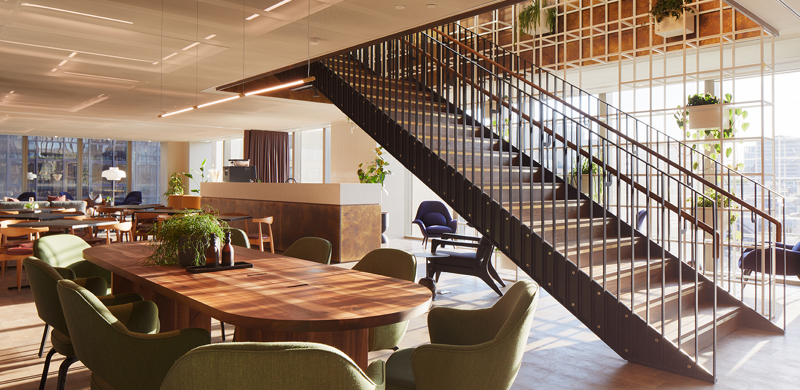 Open lounge area at MYO Liverpool Street featuring a large walnut meeting table with olive green chairs, a statement staircase with black metal detailing, lounge seating and abundant natural light.