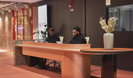 Reception area with two staff members seated behind a curved wooden desk, with dark wall panelling, pendant lights and a vase on a side table.