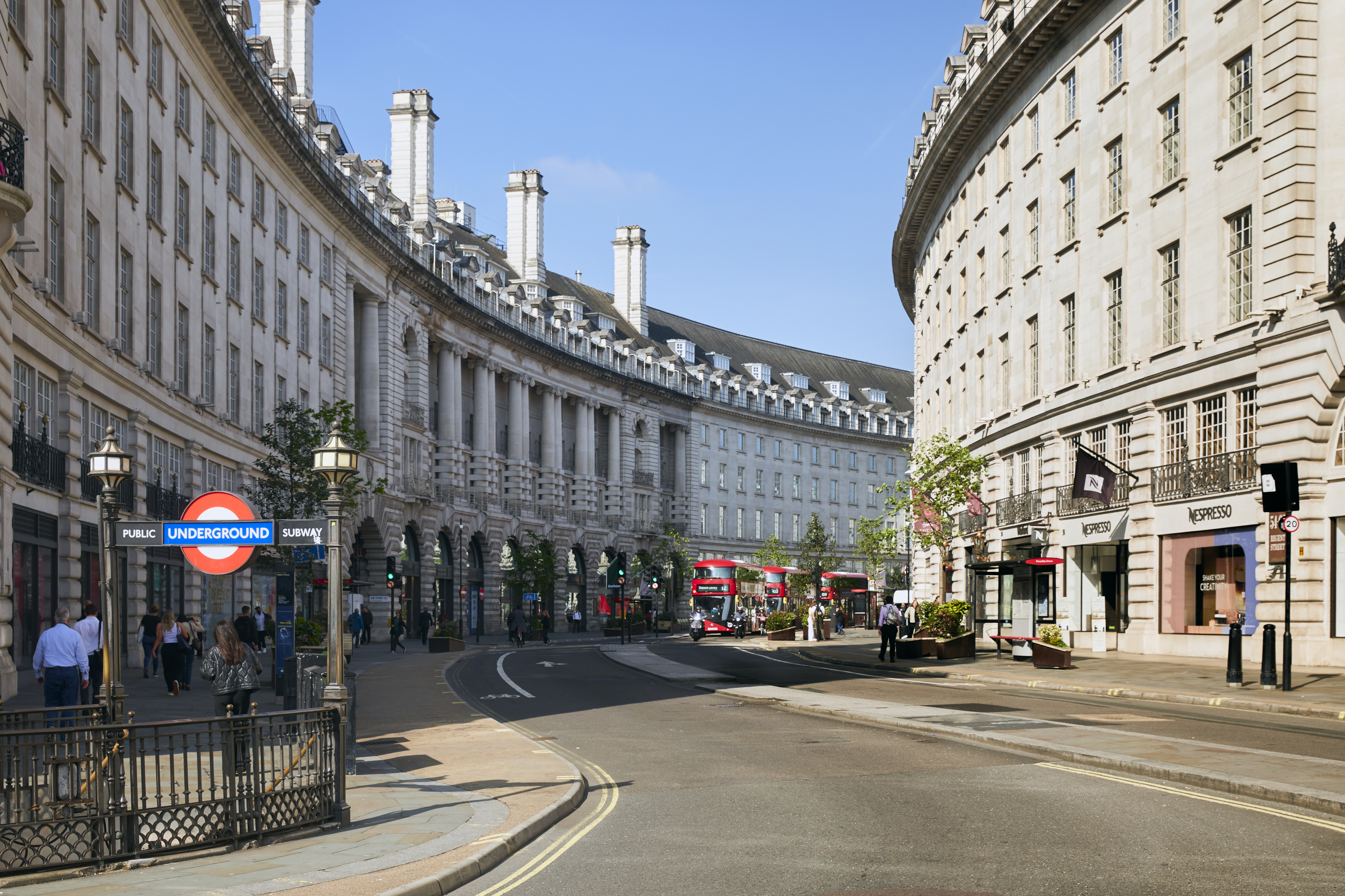 View of Regent Street in London featuring the Underground entrance, historic curved buildings, shops and a red double-decker bus on a sunny day.