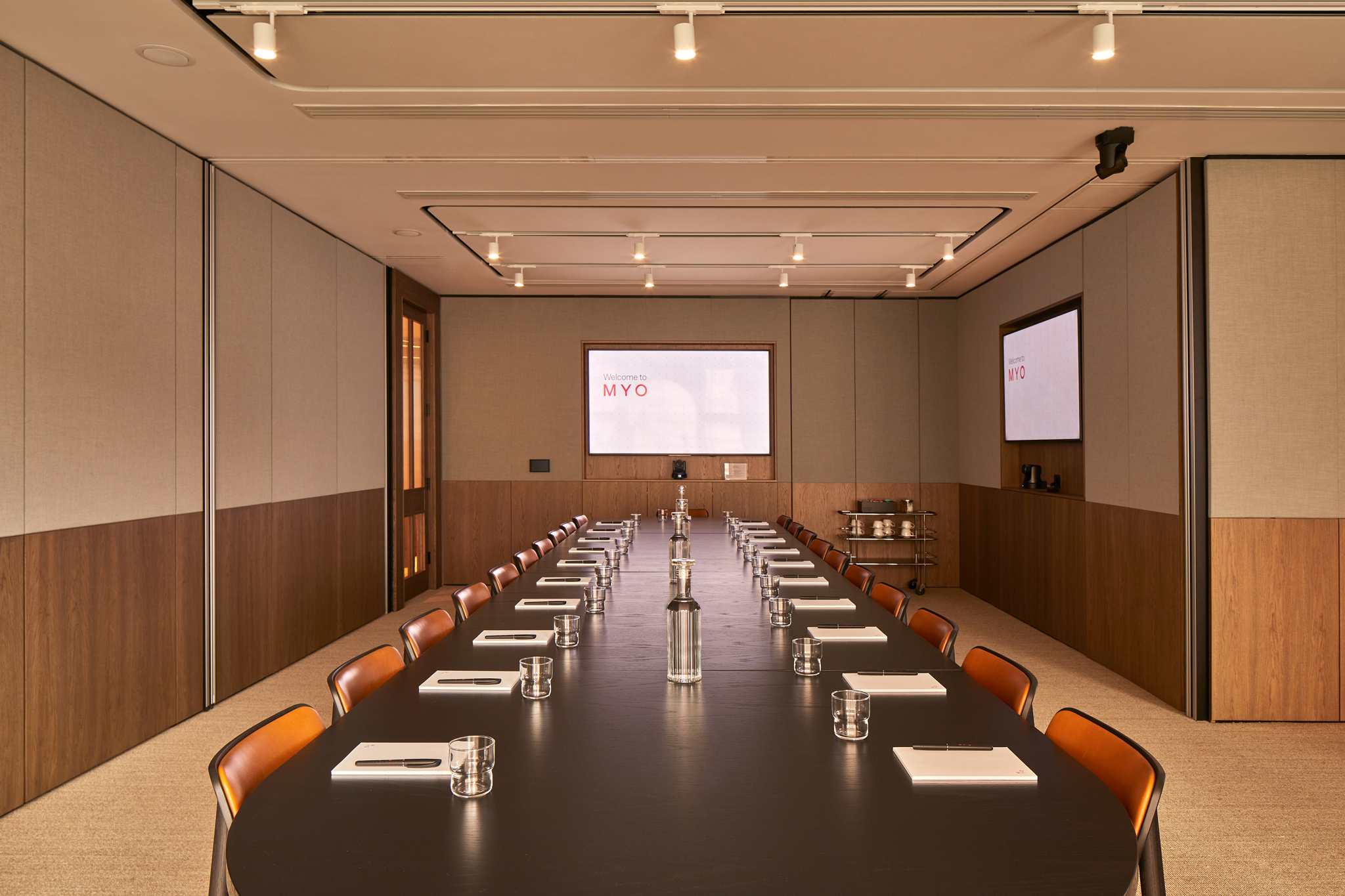 A contemporary executive boardroom featuring a long conference table, leather chairs, dual presentation screens and modern wood-panelled walls.