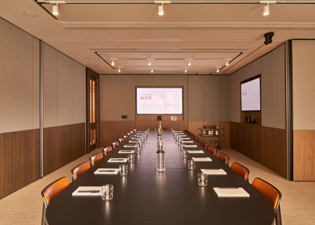 A contemporary executive boardroom featuring a long conference table, leather chairs, dual presentation screens and modern wood-panelled walls.