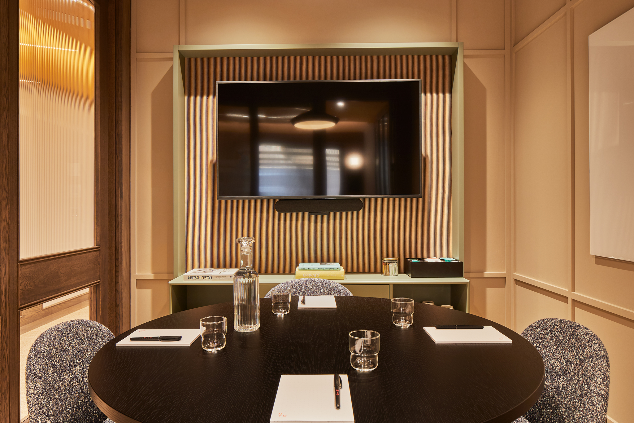 A contemporary meeting room featuring a round conference table, upholstered chairs, a wall-mounted presentation screen, integrated video conferencing equipment and warm timber finishes.