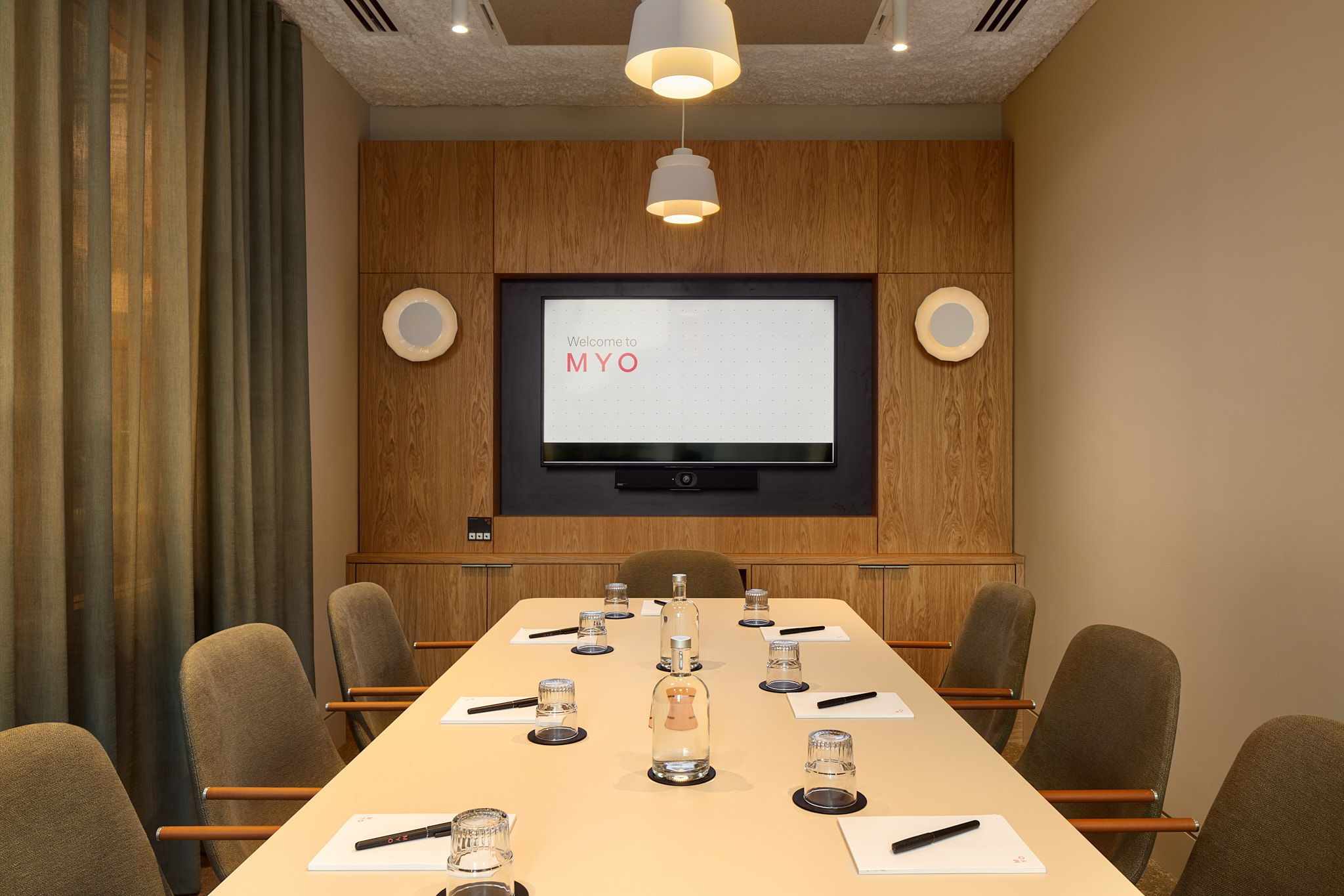 A contemporary meeting room featuring a conference table, upholstered chairs, a presentation screen, timber wall panelling and pendant lighting.