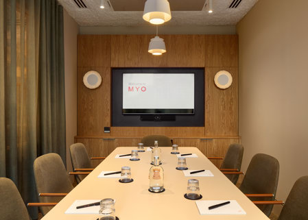 A contemporary meeting room featuring a conference table, upholstered chairs, a presentation screen, timber wall panelling and pendant lighting.