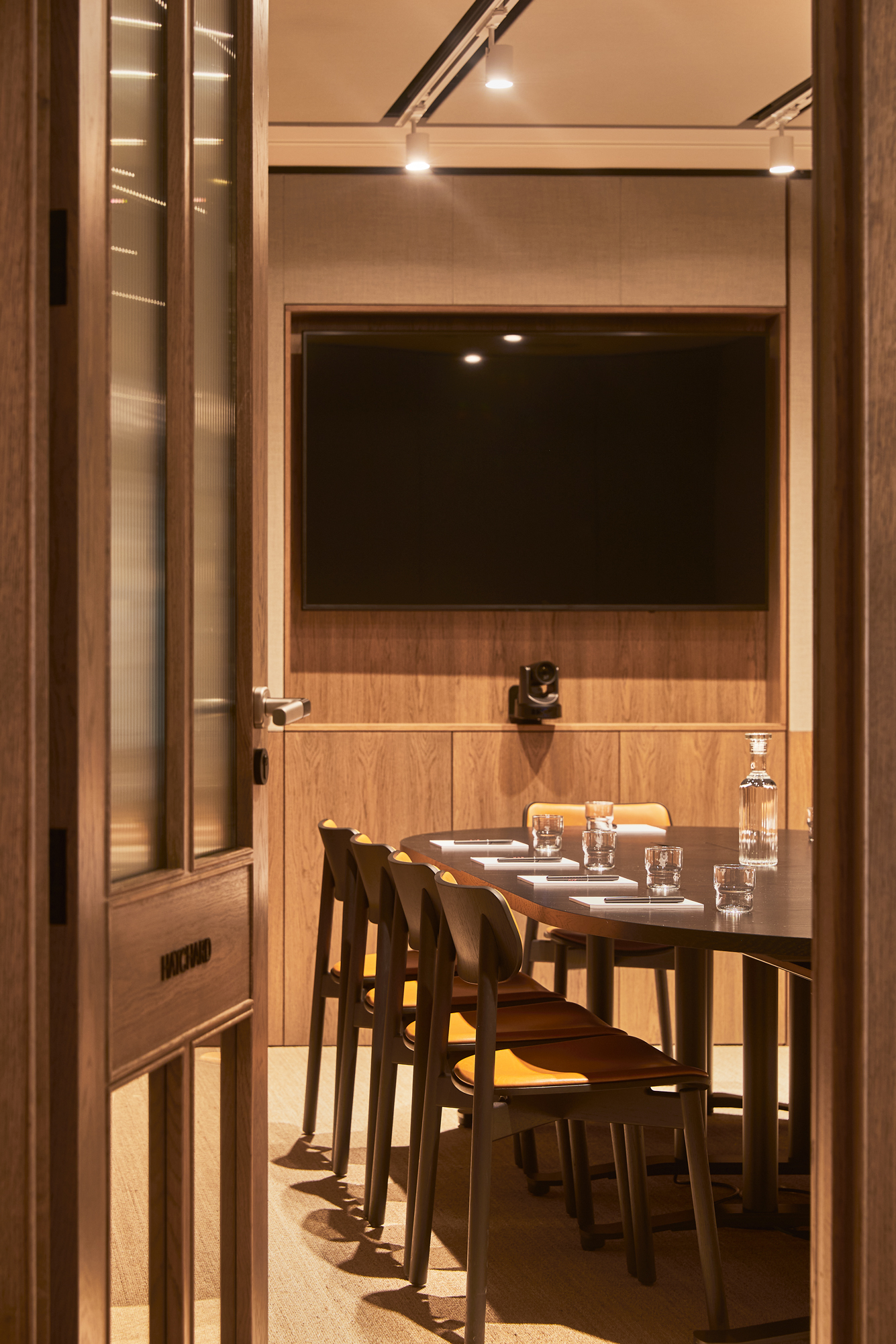 A modern meeting room with a conference table, chairs, video conferencing equipment and a wall-mounted display, viewed through an open doorway.