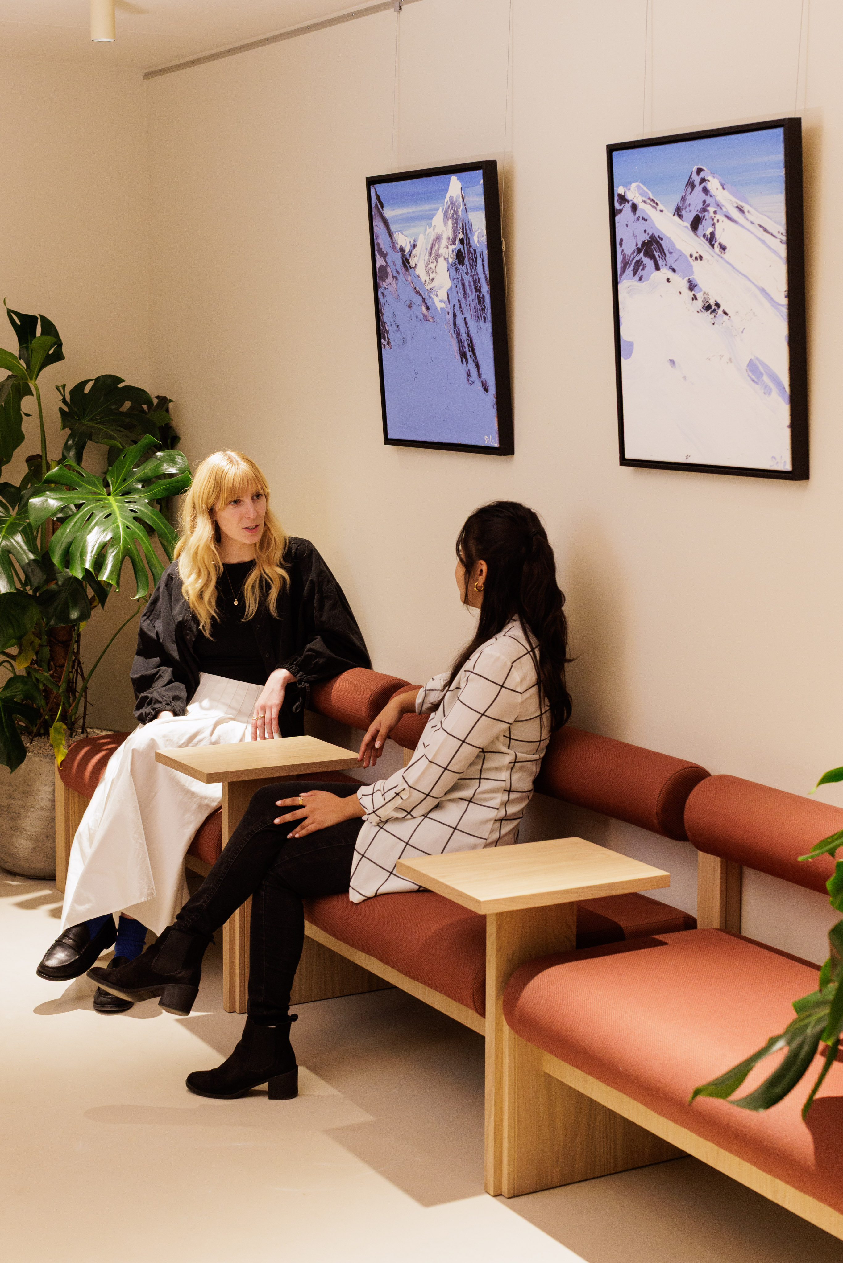 Two people having an informal meeting on upholstered bench seating in a contemporary workspace with indoor plants and framed artwork.