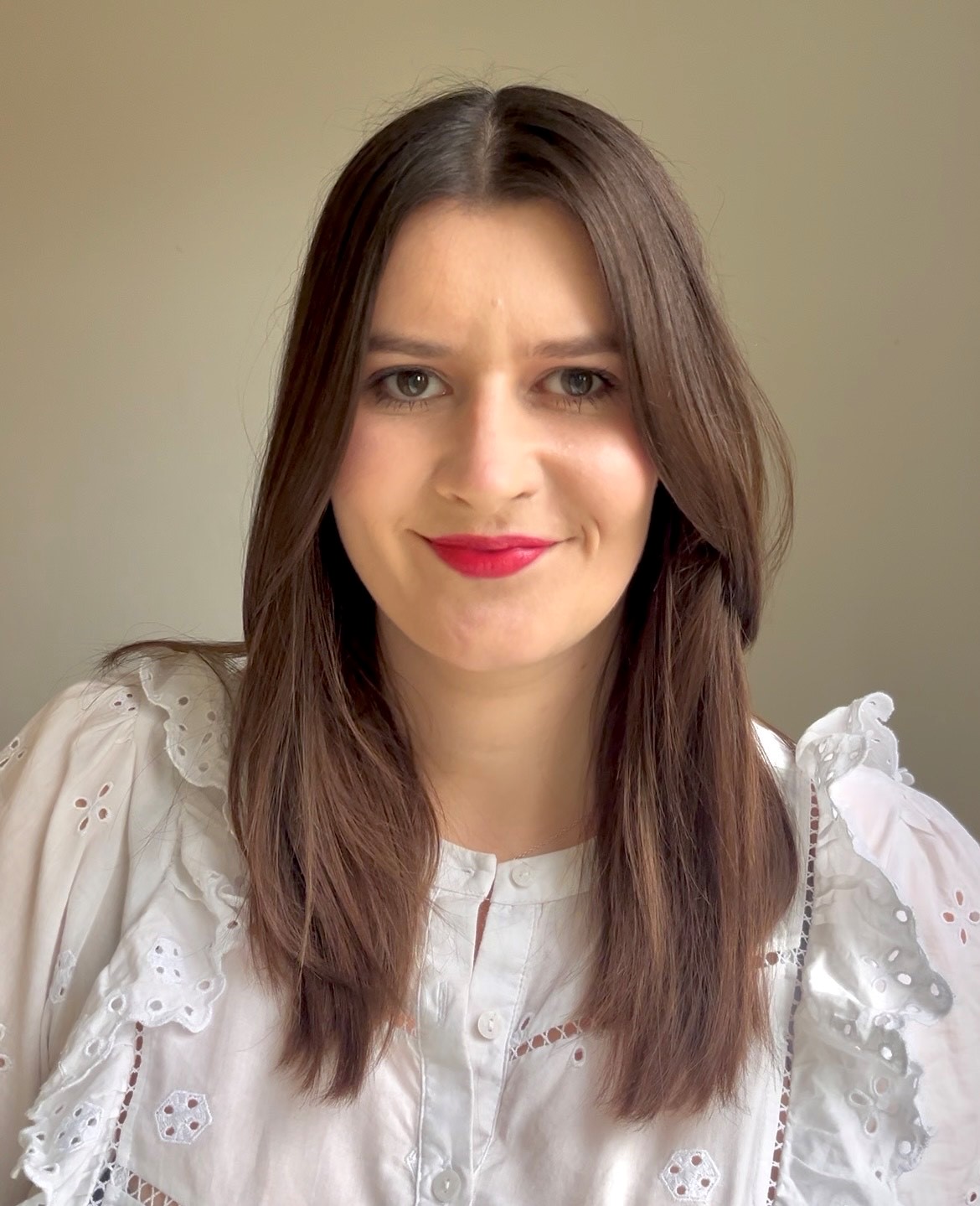 Professional headshot of a woman with shoulder-length dark brown hair wearing a white embroidered blouse with broderie detailing, photographed against a soft neutral background.