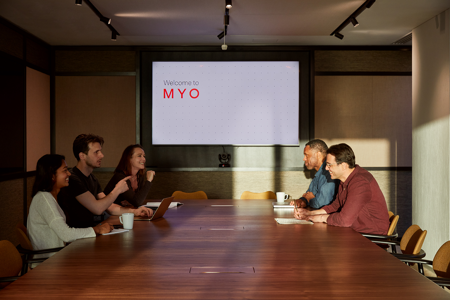 Team meeting taking place in a contemporary boardroom at MYO Liverpool Street, featuring a large walnut conference table, integrated presentation screen and warm, ambient lighting.