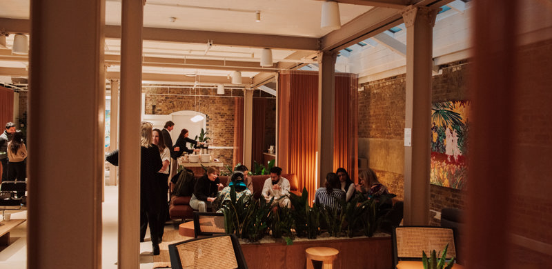 A contemporary communal lounge with people socialising, comfortable seating, indoor planting and exposed brick walls in a modern workspace.