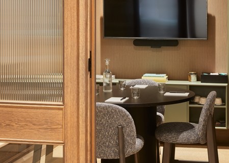 A contemporary meeting room viewed through a glazed timber door, featuring a round meeting table, upholstered chairs, a wall-mounted presentation screen and integrated video conferencing equipment.