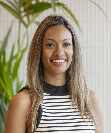 Professional headshot of a smiling woman with long highlighted hair wearing a black and white striped sleeveless top, standing in front of a softly blurred background with green planting and textured wall panels.