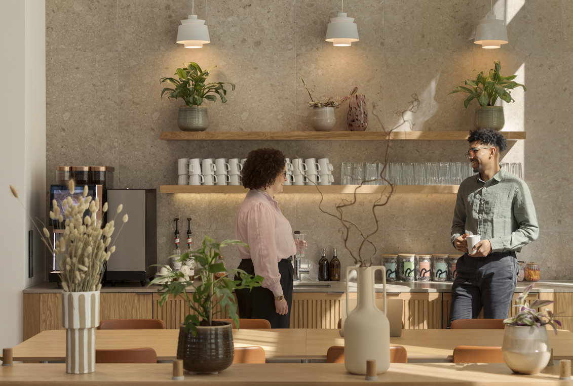 A contemporary communal kitchen featuring a coffee station, open shelving, indoor plants and two people chatting in a modern workspace.