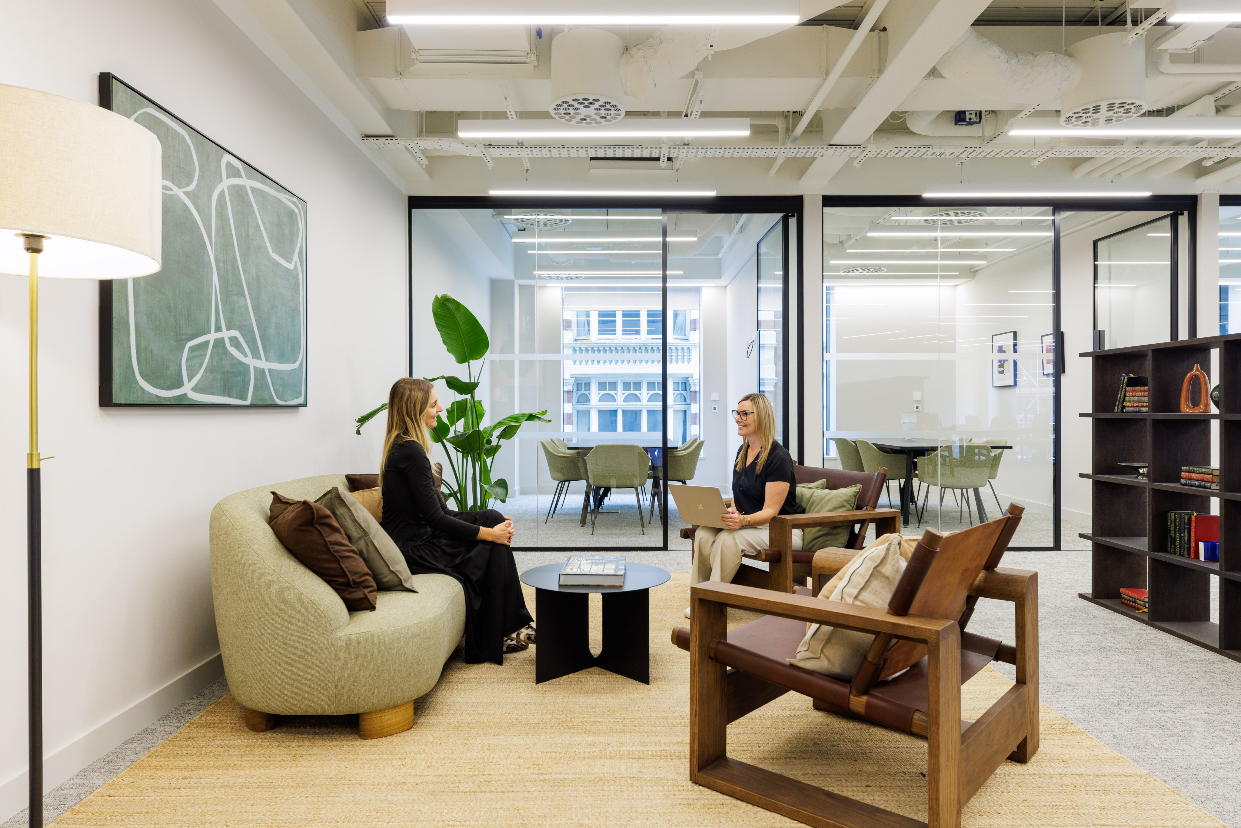 A modern lounge area with comfortable seating where two people are meeting, set beside glass meeting rooms in a contemporary office.