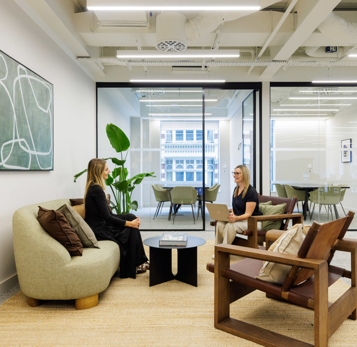 A modern lounge area with comfortable seating where two people are meeting, set beside glass meeting rooms in a contemporary office.
