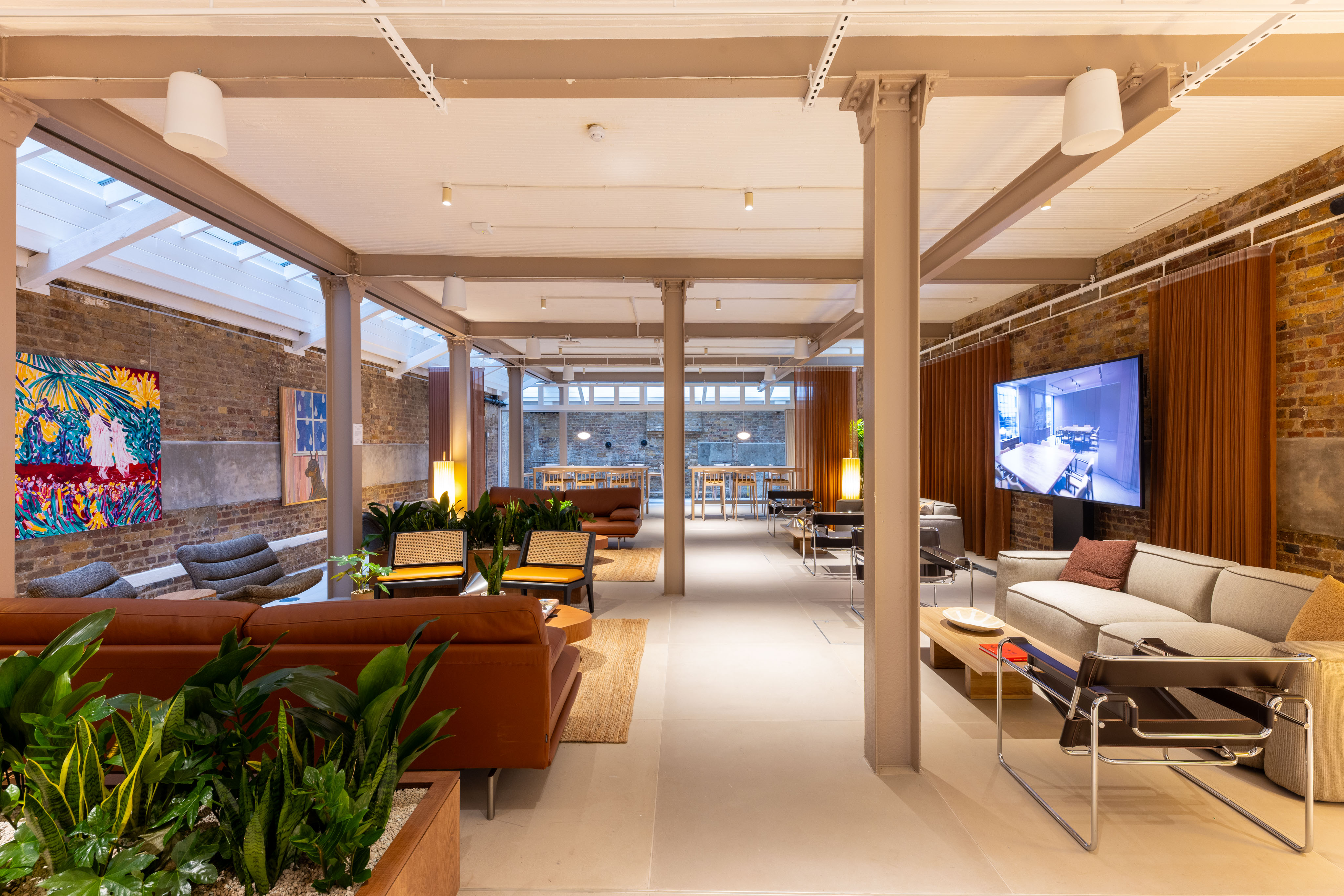 Spacious lounge-style workspace with sofas, armchairs, indoor plants, exposed brick walls, colourful artwork and a wall-mounted screen.