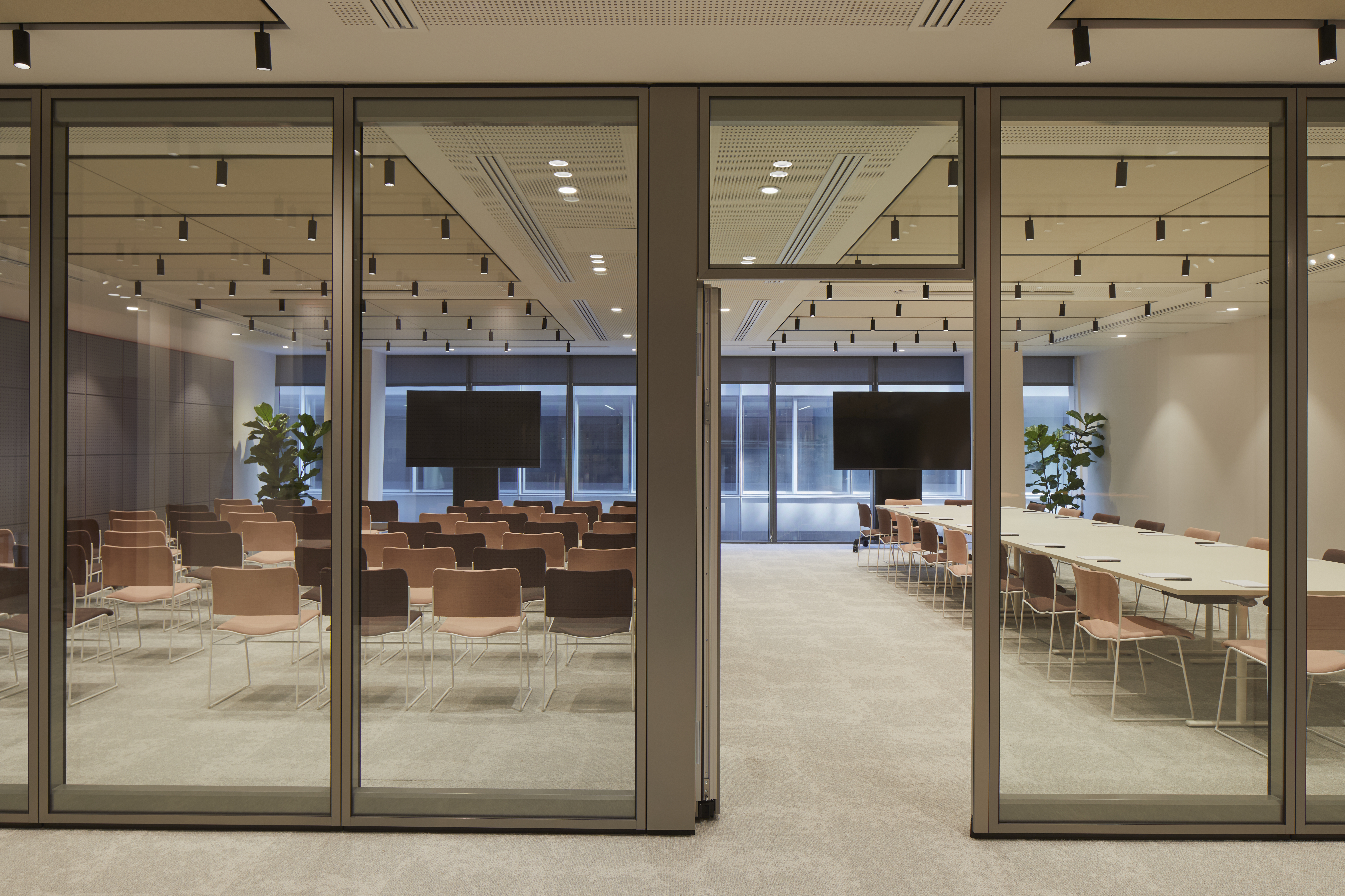 View through glass doors into a meeting space with rows of chairs, boardroom tables, large screens and floor-to-ceiling windows.