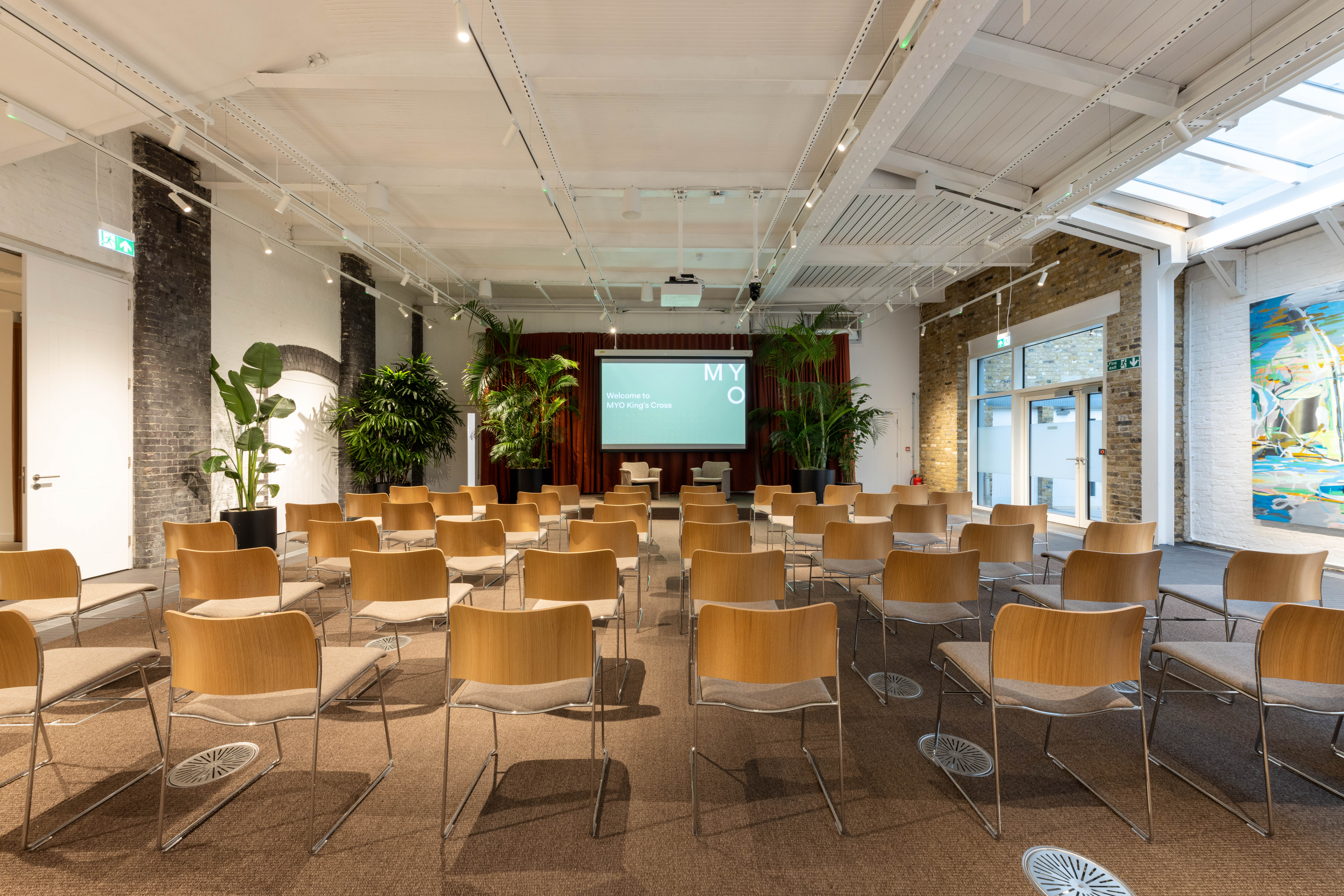 A contemporary event space with rows of chairs facing a presentation screen, surrounded by indoor plants, exposed brick walls and modern lighting.