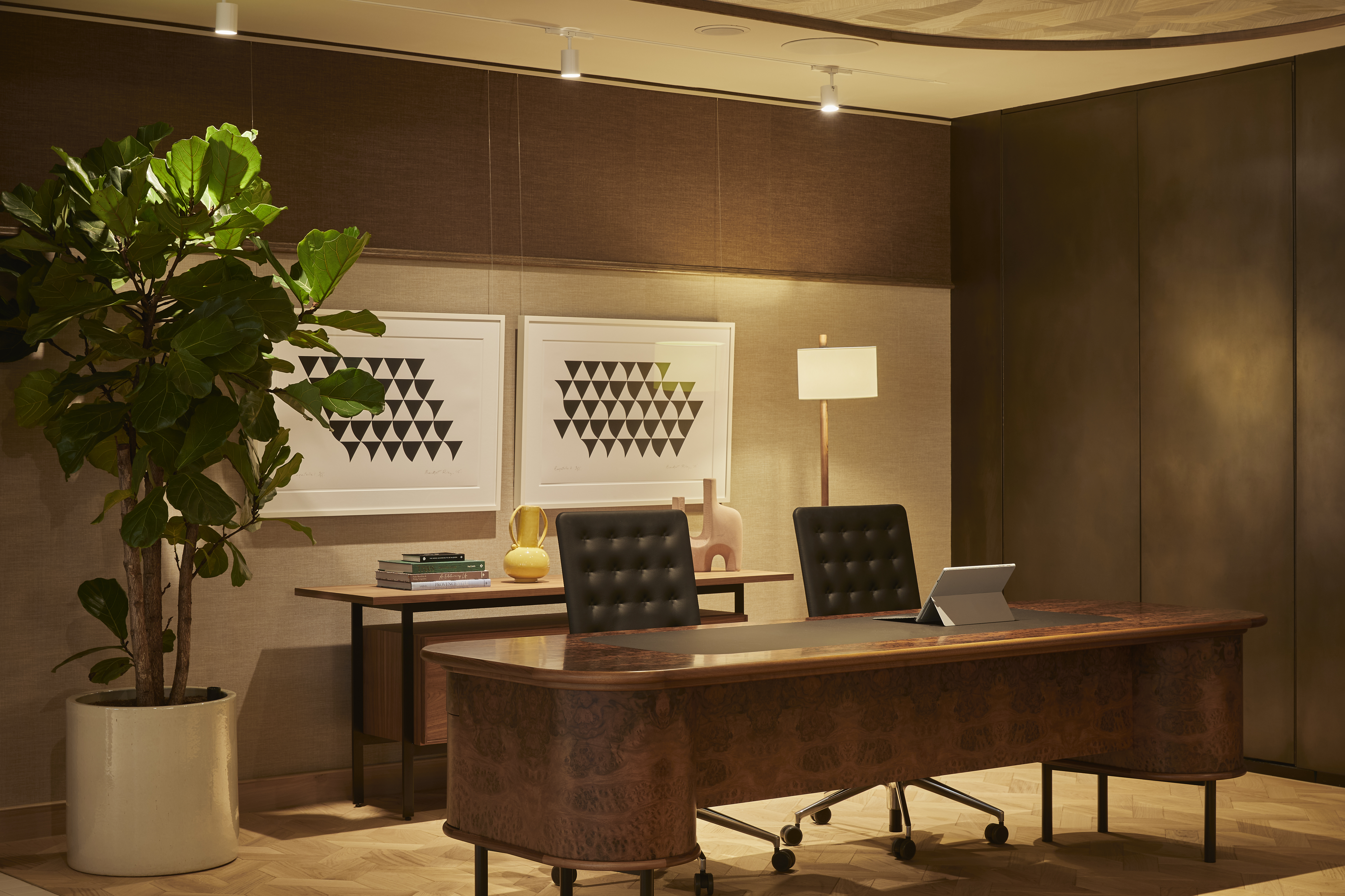 A contemporary private office featuring a wooden executive desk, office chairs, contemporary artwork, a floor lamp and a large indoor plant.