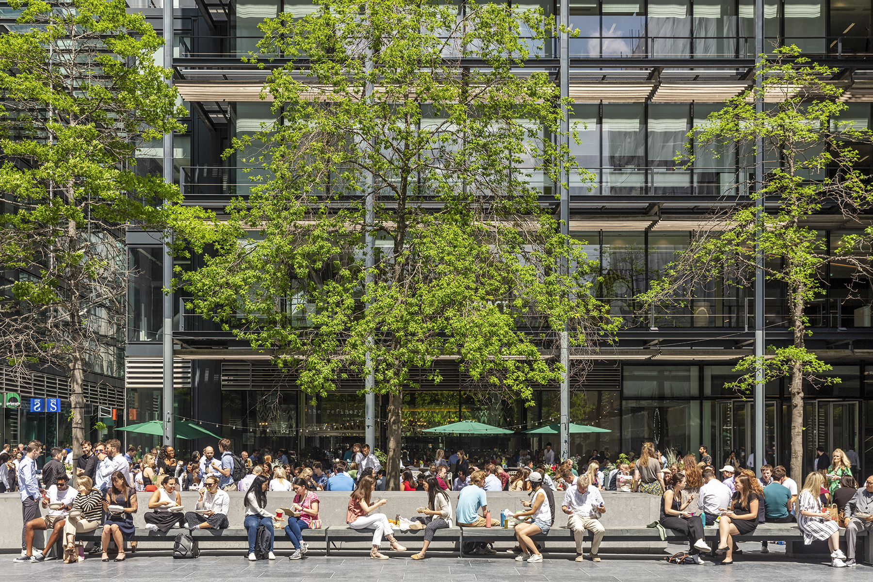 People enjoying New Street Square's outdoor plaza, with tree-lined seating areas surrounded by contemporary office buildings.