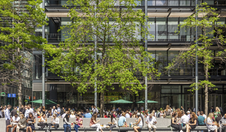 People enjoying New Street Square's outdoor plaza, with tree-lined seating areas surrounded by contemporary office buildings.