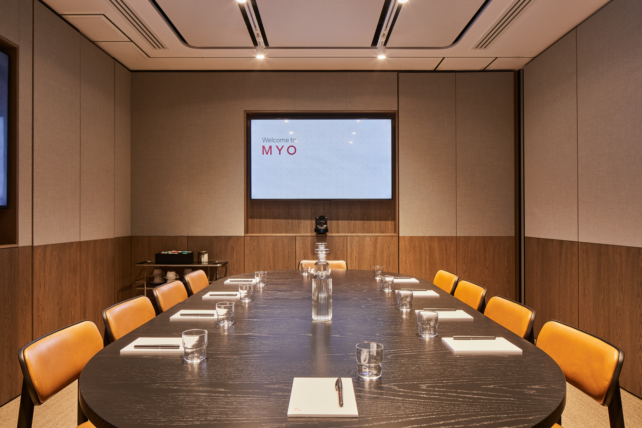 A contemporary boardroom featuring a large oval conference table, leather chairs, a presentation screen, video conferencing equipment and modern wood-panelled walls.