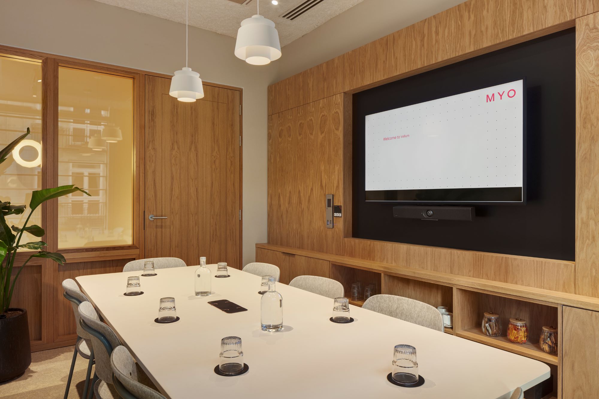 A contemporary meeting room featuring a conference table, upholstered chairs, a wall-mounted presentation screen, timber wall panelling and pendant lighting.